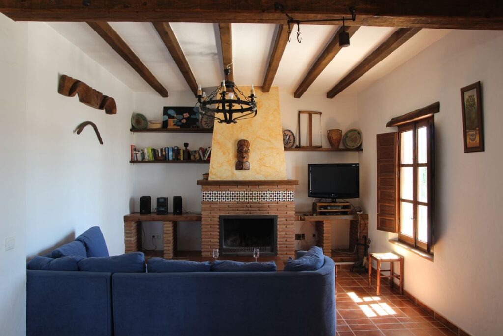 Salón en casa de campo con techo de vigas de madera y lámpara y suelo de estilo rústico, gran ventana luminosa, chimenea al fondo, TV de plasma y objetos rústicos decorando la estancia y sofá rinconera azul.