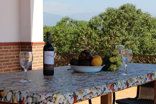 Mesa de piedra y madera con botella de vino, copas y frutero, en porche de casa rural. Constructora Málaga